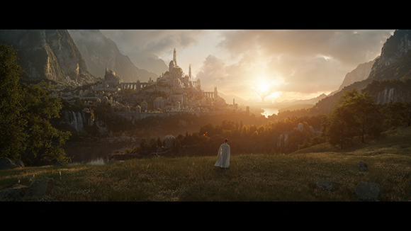 A person in a white cloak stands in a meadow looking toward a large fantasy city in a mountain valley at sunset.
