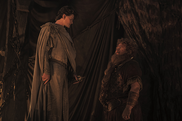 A tall figure in a light cloak looks down at a bearded man in dark armor inside a dark tent.