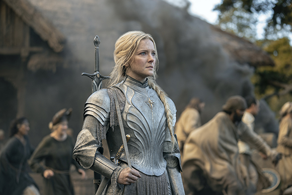 A blonde woman in silver plate armor stands in a village as smoke rises behind her.