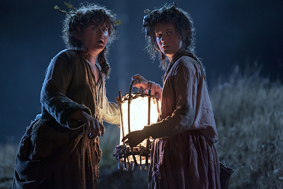 Two children in rustic clothing hold a glowing lantern in a dark, grassy field at night.