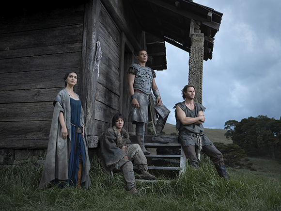 Four people in period clothing pose outside a weathered wooden structure under a cloudy sky.