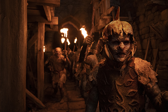 Orcs with textured skin and armor march through a dark, torch-lit stone tunnel.