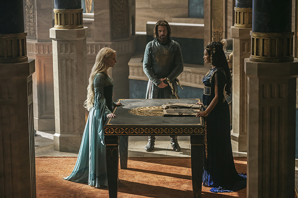 A woman in a light blue gown, a man in grey armor, and a woman in a dark blue dress stand around a table.