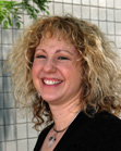 A smiling woman with curly blonde hair wearing a black top and a necklace.