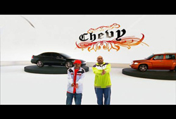 Two men stand in a white studio with a Chevy logo and two cars on platforms.
