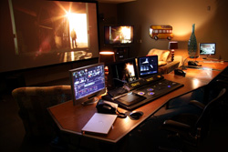 A dark editing studio featuring a large projection screen, several computer monitors, and a long wooden desk.
