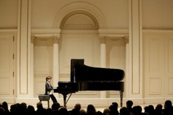 A young person sits at a black grand piano on a large, formal stage in front of an audience.