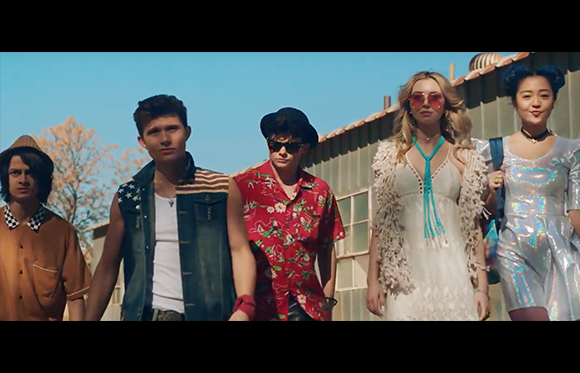 Five young people in stylish, colorful clothing stand side-by-side against a building and blue sky.
