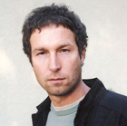 A man with dark hair and light stubble looking forward against a plain background.