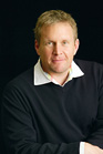 A man with short blond hair wearing a black sweater over a white collared shirt against a black background.