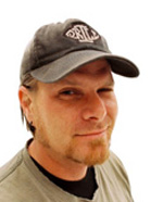 A man with a goatee and a grey baseball cap smiling against a white background.