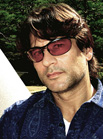 A man with dark hair wearing reddish-tinted sunglasses and a blue patterned shirt.