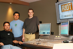 Three men posing in a recording studio with a mixing console and monitors.