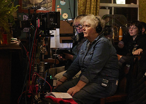 A woman in a denim jacket wearing headphones sits near a professional film camera and crew members.