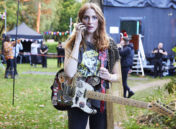 A woman with long red hair holds a white electric guitar and talks on a mobile phone outdoors.