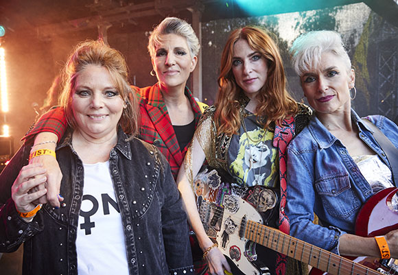 Four women in stylish, eclectic clothing posing closely together in a brightly lit setting.