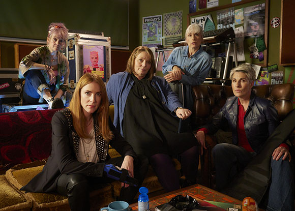 Five women sit and stand in a room decorated with posters and musical equipment.