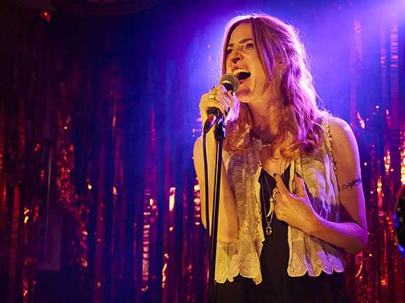 A woman with long reddish hair sings into a microphone under purple stage lighting.