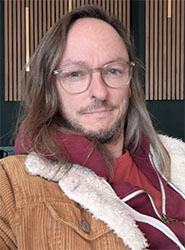 A man with long brown hair and glasses wearing a maroon hoodie and a brown corduroy jacket.
