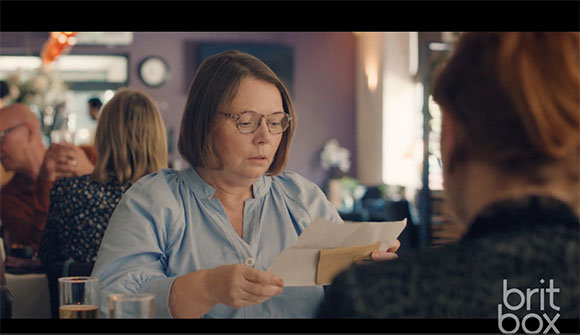 A woman with glasses sits at a table, looking down intently at an open envelope and paper.