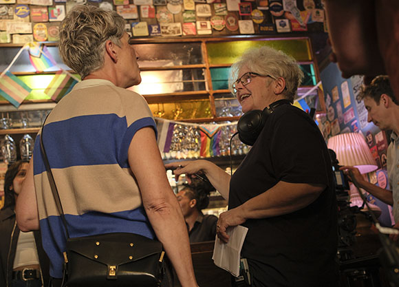 Two women engage in conversation in a room filled with various objects and shelves.