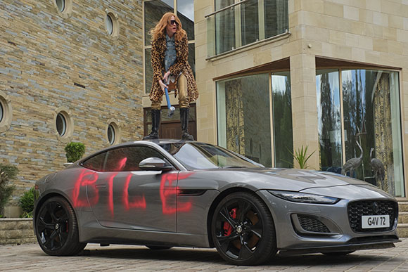 A person in a leopard print coat stands behind a grey sports car with red graffiti on the side.