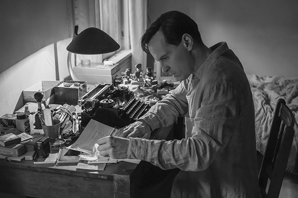 A man in a button-down shirt sits at a desk covered in papers and a typewriter, writing on a document.