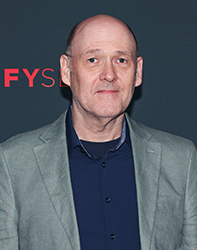 A bald man wearing a grey blazer over a dark blue button-down shirt against a dark background.