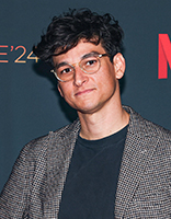 A man with dark curly hair and glasses wearing a grey patterned blazer and black t-shirt.