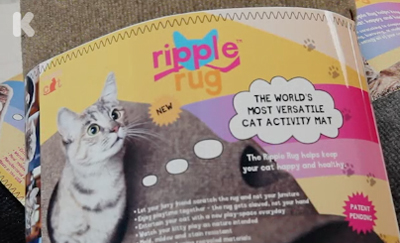 A magazine advertisement for the Ripple Rug cat activity mat featuring a photo of a grey and white cat.