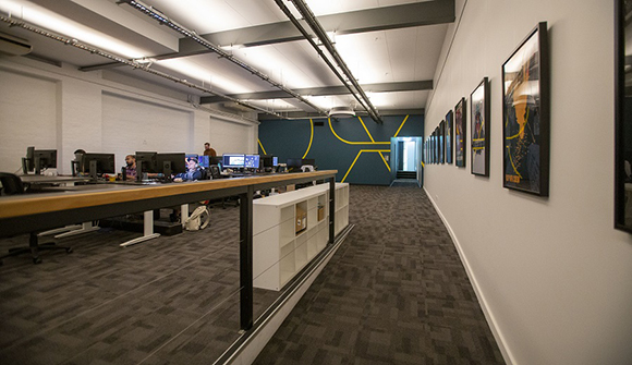 An open office with long wooden desks, computer monitors, and framed artwork on a white wall.