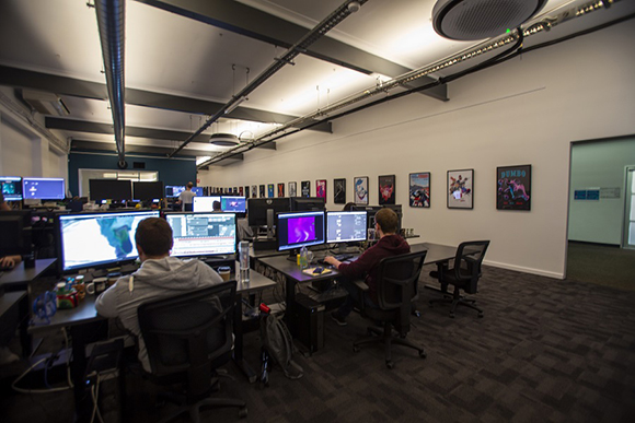 People sit at desks with multiple monitors in a brightly lit, open-plan office with art on the walls.