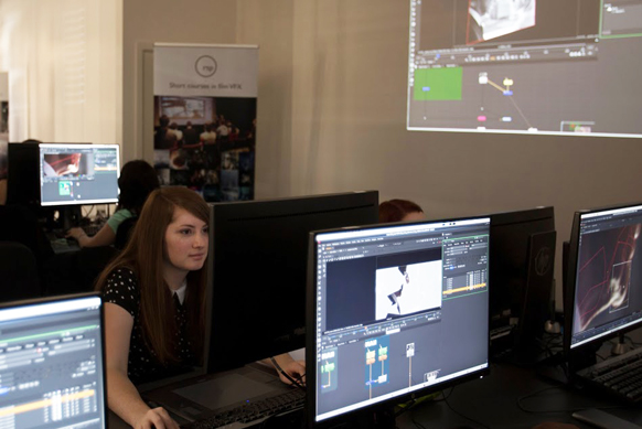 A young woman sits at a desk using a computer in a classroom filled with monitors and digital displays.