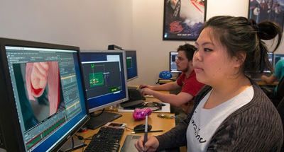 A woman uses a stylus on a tablet to edit digital artwork on a computer monitor in a classroom setting.