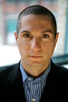 A man with short dark hair wearing a blue striped shirt and a black blazer looks directly at the camera.