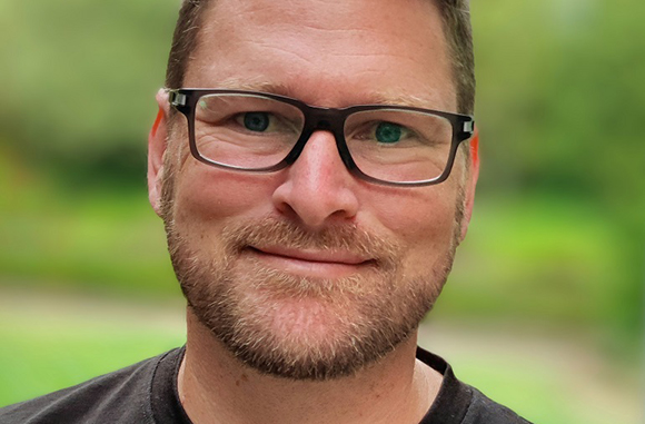 Close-up portrait of a man with short brown hair, glasses, and a beard smiling against a blurred green background.