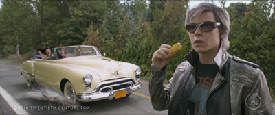A young man in a leather jacket and sunglasses holds a piece of fruit with a yellow convertible driving behind him.