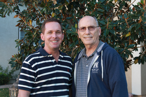 Two men, one younger in a striped polo and one older in a blue hoodie, smiling in front of a large green bush.
