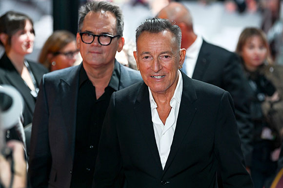Two men in dark suits and button-down shirts walking through a crowd.