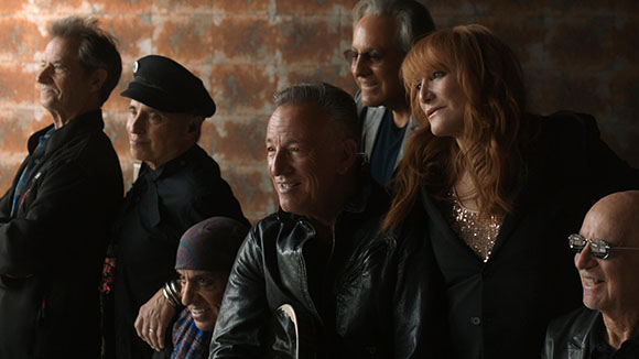 A group of five people in dark clothing looking in various directions against a textured wall.
