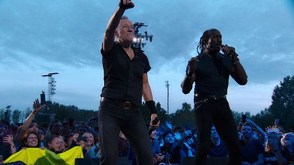 Two male singers perform on an outdoor stage in front of a large crowd under a blue twilight sky.