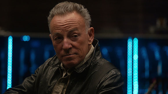 A man with short hair wearing a black leather jacket looks toward the camera against a dark, blue-lit background.