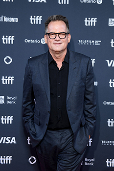 A man wearing a dark suit, black shirt, and glasses poses against a blue TIFF branded backdrop.