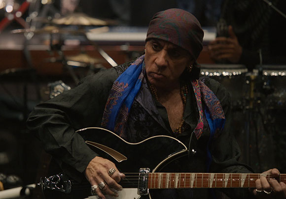 A person with a patterned headscarf plays a black electric guitar in a dimly lit setting.
