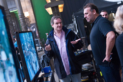 A man in a dark jacket gestures with both hands while talking to a man in a black polo shirt.