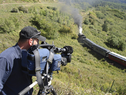 A man in a black cap uses a professional video camera on a tripod to film a steam train in a hilly landscape.