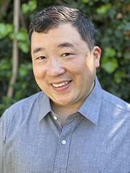 A smiling man with short dark hair wearing a grey button-down shirt against a background of green leaves.