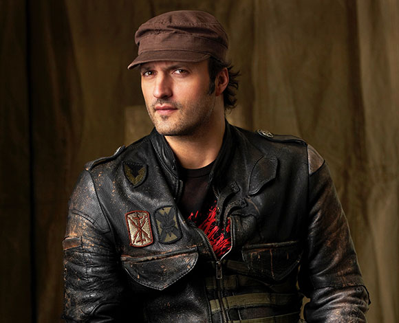 A man wearing a brown cap and a distressed black leather jacket against a dark background.