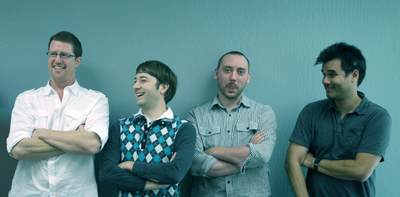 Four men standing side by side against a solid blue background.