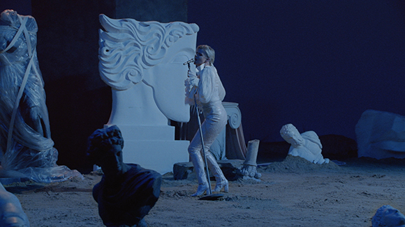 A person in a white outfit sings into a microphone amidst large white sculptures on a dark stage.